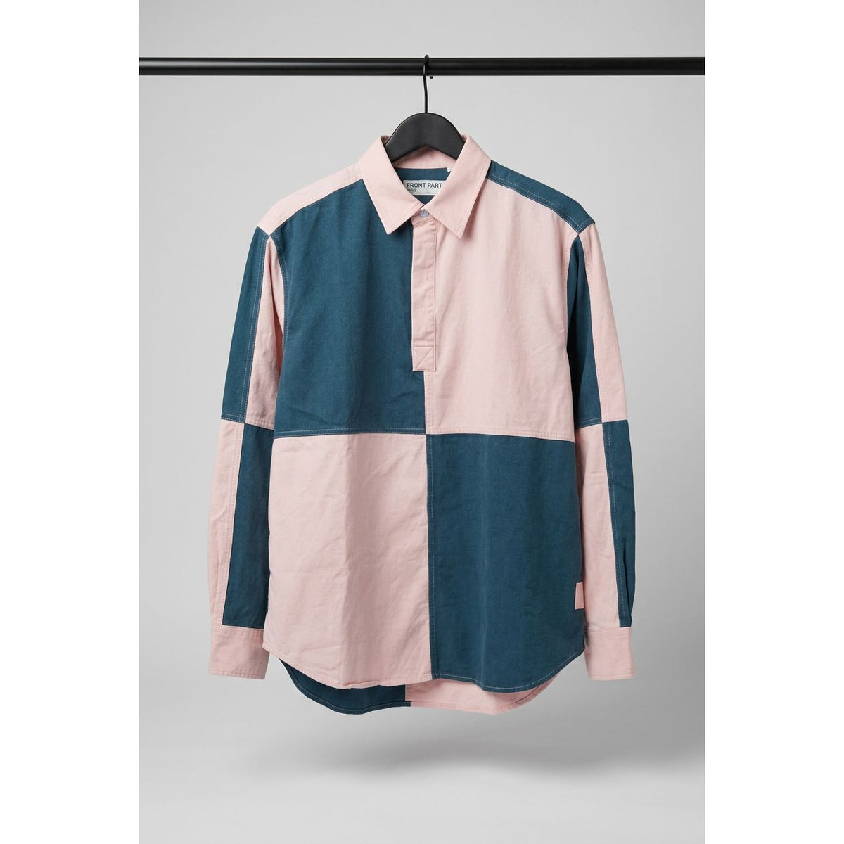 A long-sleeve colorblock shirt with a pink collar and button placket, featuring large blocks of dark teal blue and light pink, hanging on a black hanger against a wooden door.