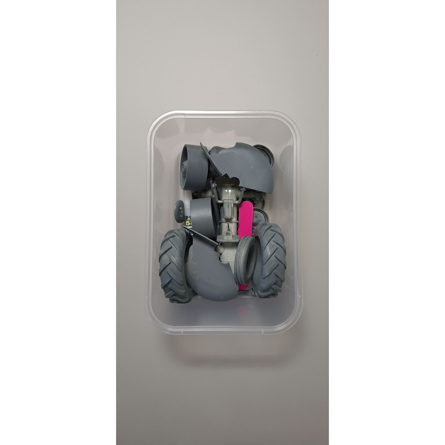 Toy robot with large wheels and a pink component inside a transparent container on a beige surface.